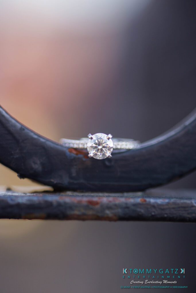 JamieMark-Engagement-11 - TOMMY GATZ ENTERTAINMENT A diamond ring is placed on a metal surface with a blurred background.