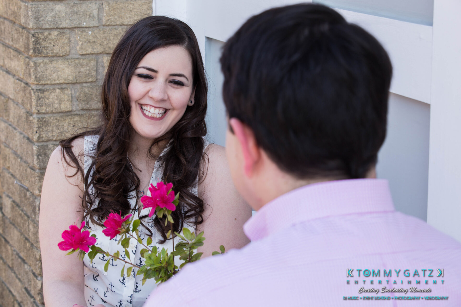 JamieMark-Engagement-15 - TOMMY GATZ ENTERTAINMENT A woman smiles while holding pink flowers, facing a man in a pink shirt. The background includes a brick wall and white paneling.