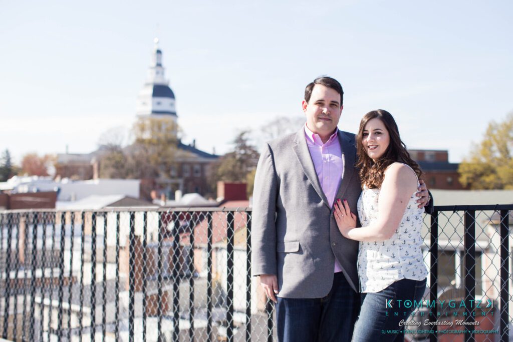 JamieMark-Engagement-18 - TOMMY GATZ ENTERTAINMENT A couple stands together on an outdoor balcony overlooking a cityscape with a domed building in the background.