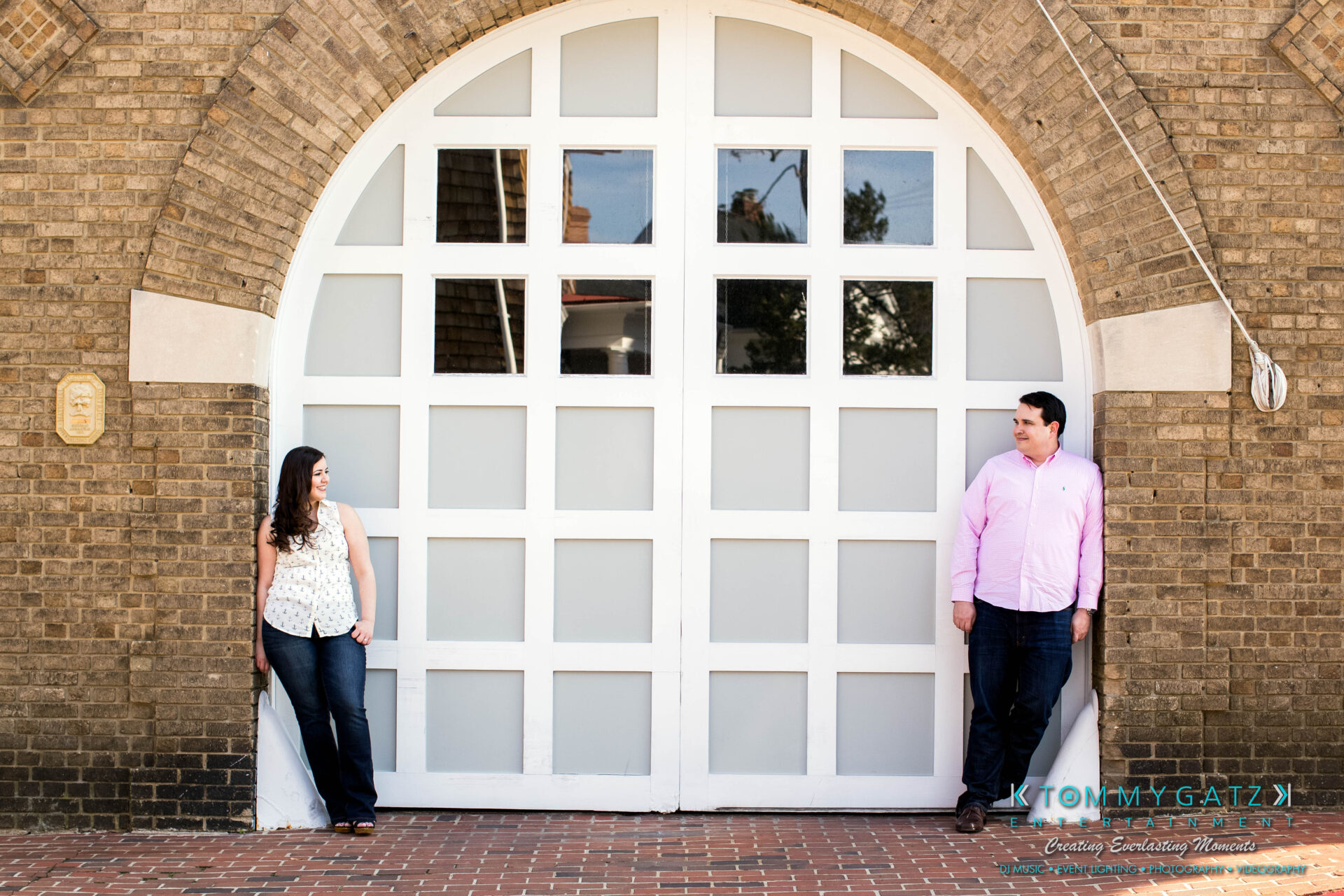 JamieMark-Engagement-19 - TOMMY GATZ ENTERTAINMENT A man and a woman stand apart, leaning against a large arched doorway with a white grid design on a brick building.