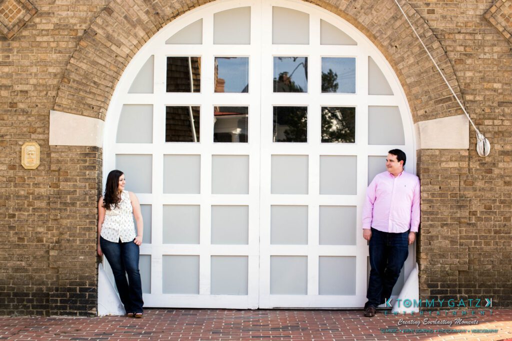 JamieMark-Engagement-19 - TOMMY GATZ ENTERTAINMENT A man and woman stand apart, leaning against a large arched white garage door on a brick building, facing each other and wearing casual outfits.