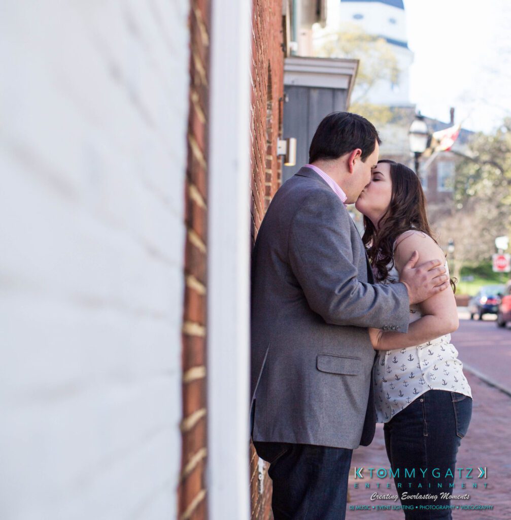 JamieMark-Engagement-2 - TOMMY GATZ ENTERTAINMENT A couple shares a kiss on a brick sidewalk, leaning against a building. The man wears a gray blazer, and the woman wears a patterned sleeveless top. Trees and a blue sky are in the background.