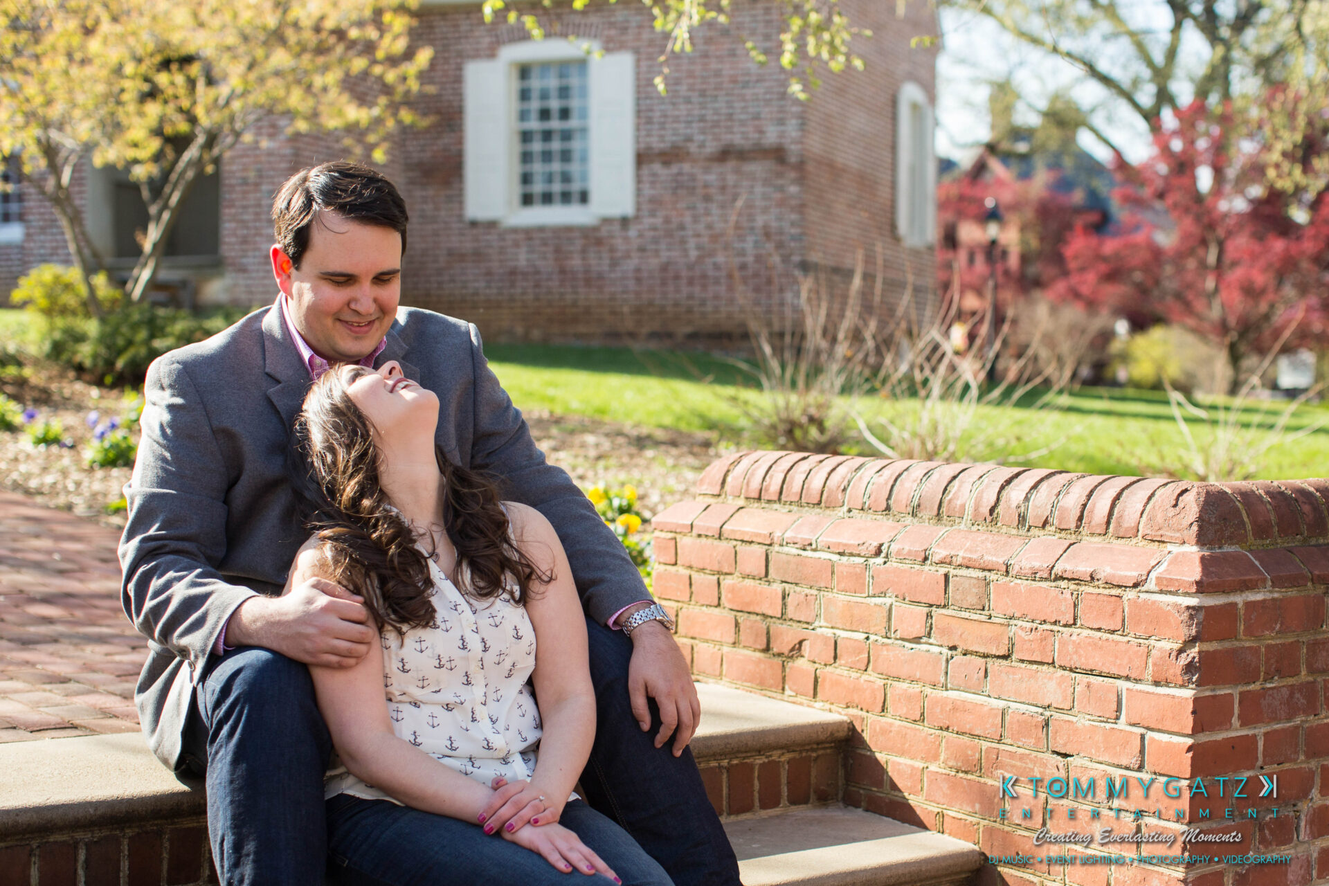 JamieMark-Engagement-4 - TOMMY GATZ ENTERTAINMENT A couple sits on brick steps outside, with the man smiling and the woman leaning back, smiling upwards.