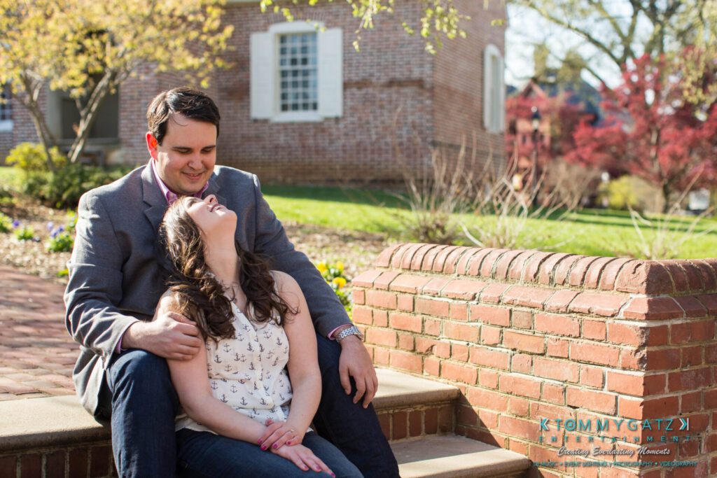 JamieMark-Engagement-4 - TOMMY GATZ ENTERTAINMENT A couple sits on steps outside, smiling and enjoying a sunny day. The background features a brick building and blooming trees.