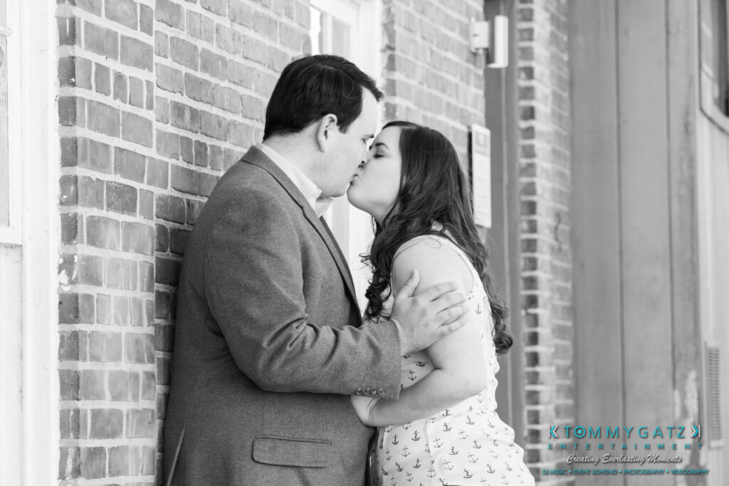JamieMark-Engagement-47 - TOMMY GATZ ENTERTAINMENT A couple kisses against a brick wall, with the man in a blazer and the woman in a patterned dress.