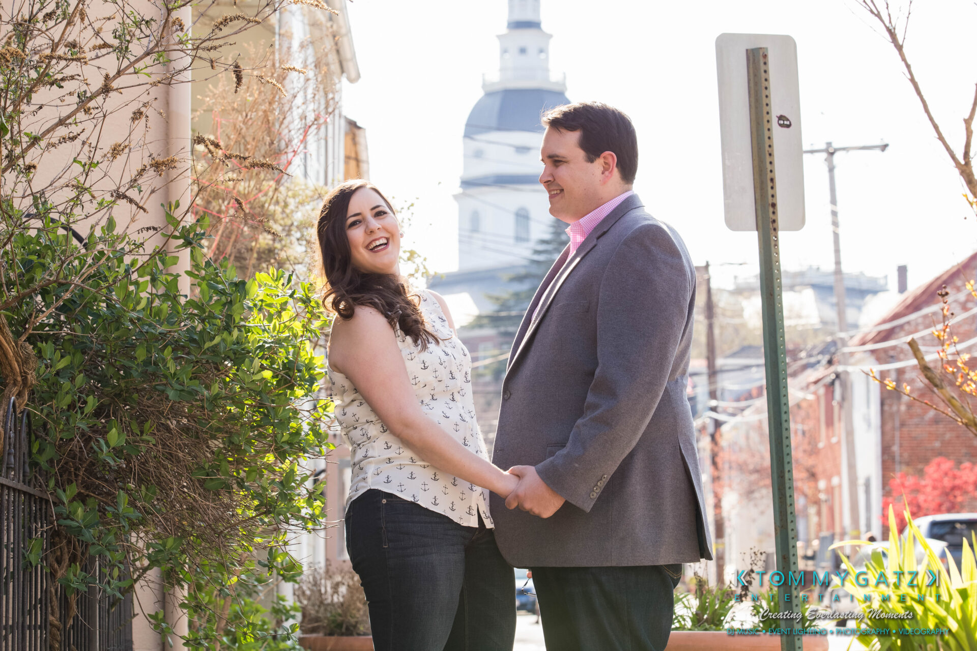 JamieMark-Engagement-49 - TOMMY GATZ ENTERTAINMENT A man and woman hold hands and smile on a sunny street with a large white building in the background.