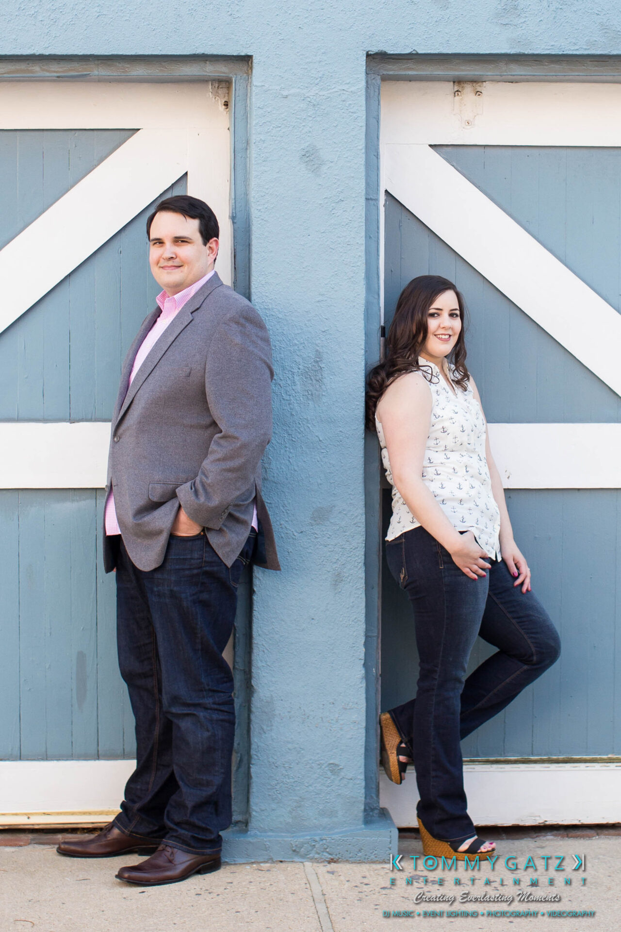JamieMark-Engagement-6 - TOMMY GATZ ENTERTAINMENT A man and woman stand back-to-back against a blue wall with white cross design. The man wears a blazer and jeans; the woman is in a patterned top and jeans. .