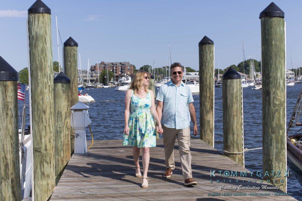 Jennifer-Rudi-Engagement-6102017-Blog-WM-1 - TOMMY GATZ ENTERTAINMENT A couple walks hand in hand on a dock by the marina, with boats and buildings visible in the background on a sunny day.