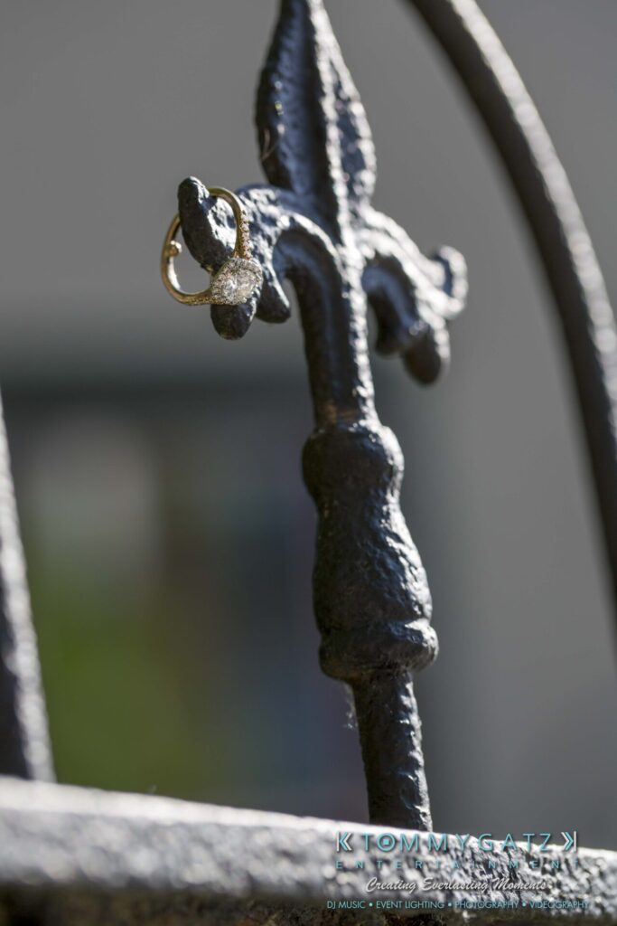 Jennifer-Rudi-Engagement-6102017-Blog-WM-10 - TOMMY GATZ ENTERTAINMENT A diamond ring hangs on a decorative metal fence post in close-up view.