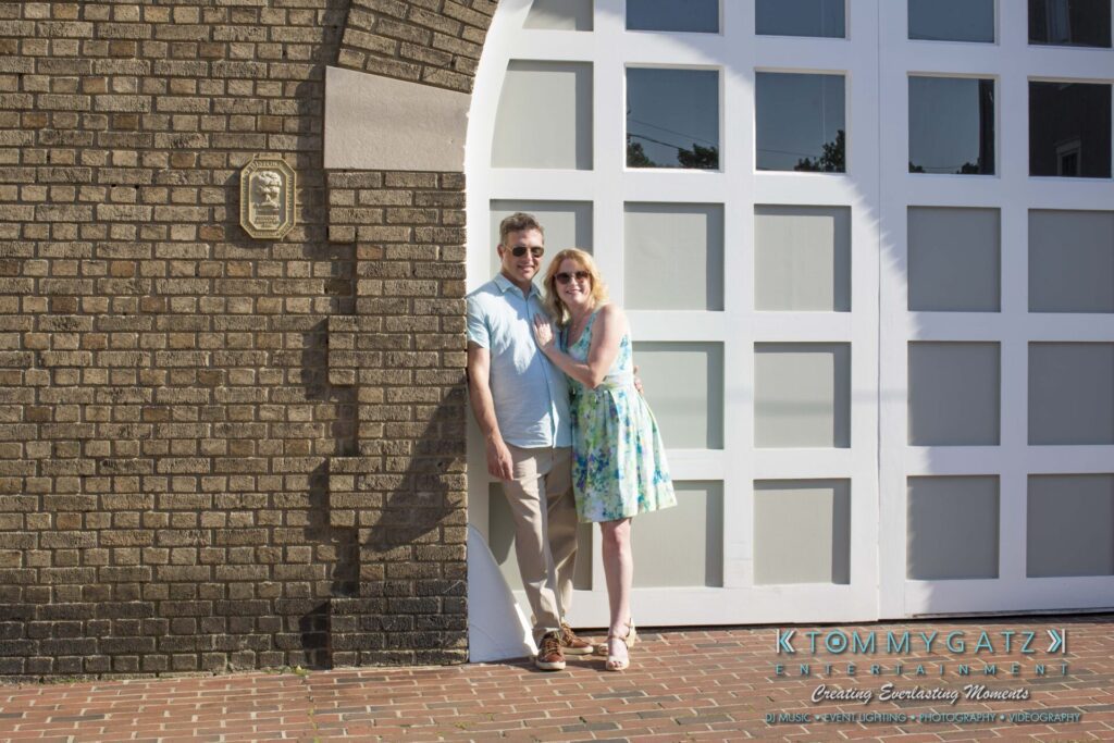 Jennifer-Rudi-Engagement-6102017-Blog-WM-12 - TOMMY GATZ ENTERTAINMENT A couple stands smiling against a brick wall next to a large white door. The man wears a light blue shirt and khaki pants, while the woman wears a blue floral dress.