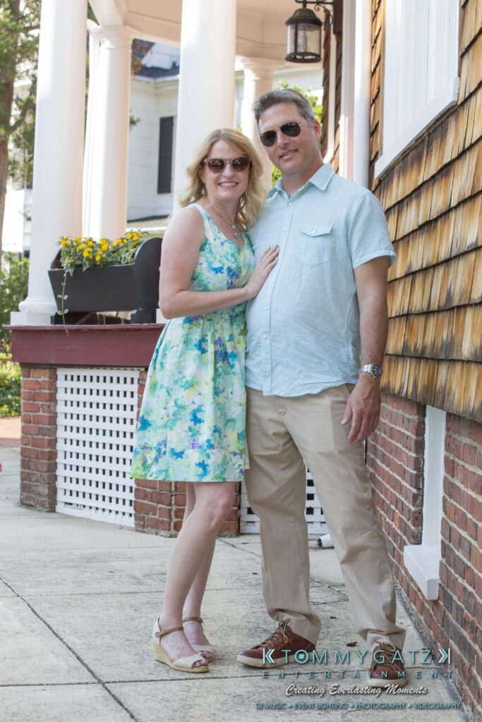 Jennifer-Rudi-Engagement-6102017-Blog-WM-16 - TOMMY GATZ ENTERTAINMENT A couple posing for a photo outside a building with columns. The woman is wearing a floral dress, and the man is in a light blue shirt and beige pants.