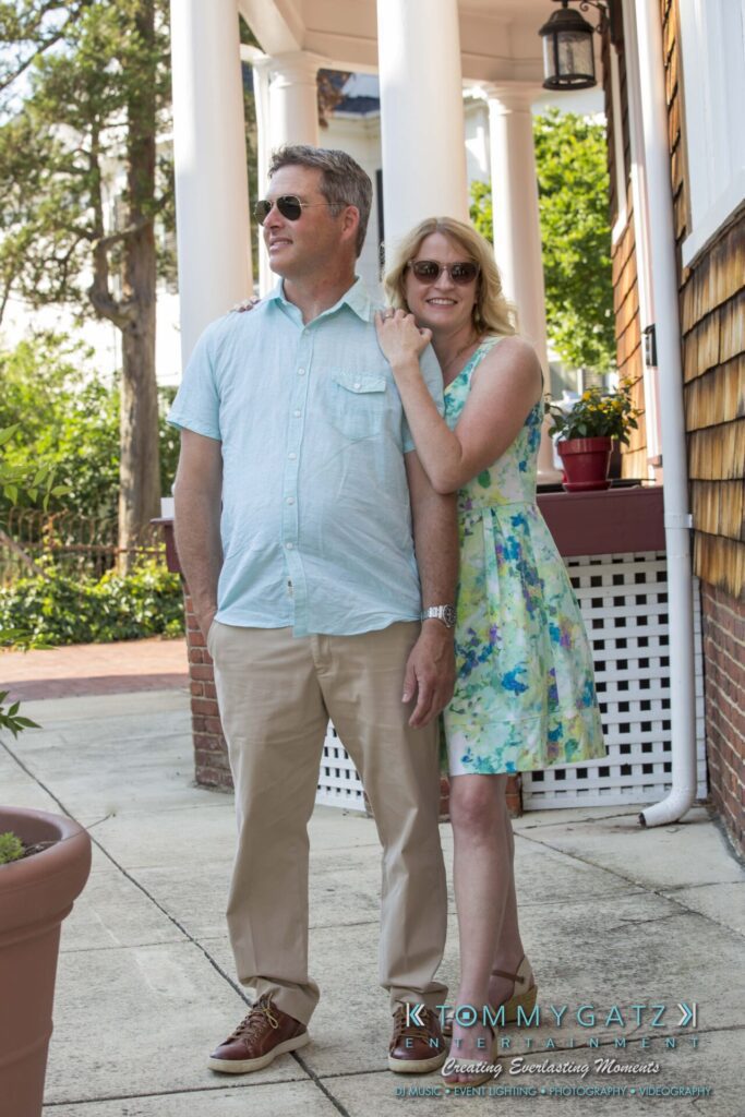 Jennifer-Rudi-Engagement-6102017-Blog-WM-18 - TOMMY GATZ ENTERTAINMENT A man and a woman stand together outdoors, smiling. The man wears a light shirt and khaki pants, and the woman wears a floral dress and sunglasses. They are on a sidewalk near a house.