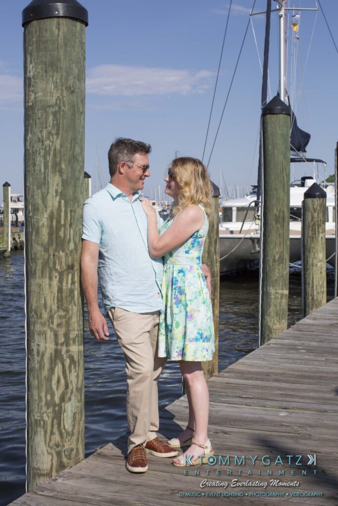 Jennifer-Rudi-Engagement-6102017-Blog-WM-2 - TOMMY GATZ ENTERTAINMENT A couple stands on a wooden dock beside sailboats, with the woman touching the man's shoulder. They're both wearing light, summery clothing under a clear blue sky.
