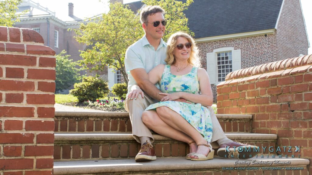 Jennifer-Rudi-Engagement-6102017-Blog-WM-27 - TOMMY GATZ ENTERTAINMENT A couple sitting together on brick steps outside, surrounded by lush greenery and sunlight.