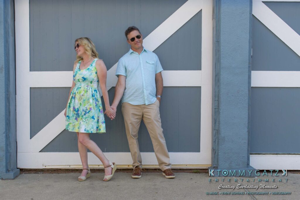 Jennifer-Rudi-Engagement-6102017-Blog-WM-3 - TOMMY GATZ ENTERTAINMENT A couple stands holding hands in front of a large blue and white barn door, both wearing sunglasses; the woman is in a floral dress and the man is in a light blue shirt and beige pants.