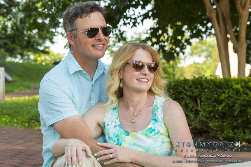 Jennifer-Rudi-Engagement-6102017-Blog-WM-30 - TOMMY GATZ ENTERTAINMENT A couple sits together outdoors on a sunny day, both wearing sunglasses.