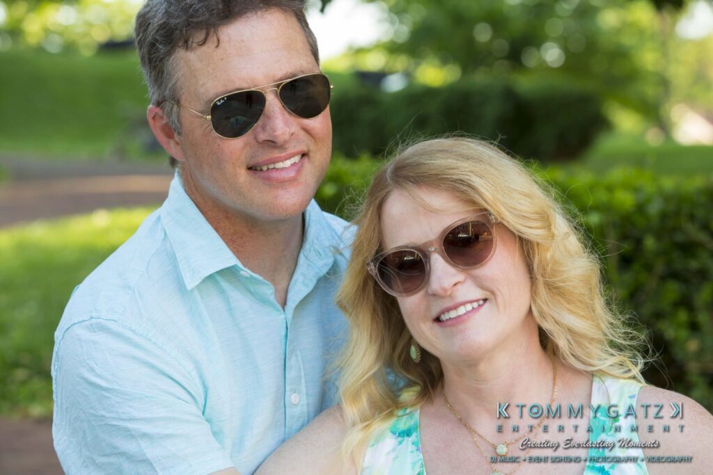 Jennifer-Rudi-Engagement-6102017-Blog-WM-31 - TOMMY GATZ ENTERTAINMENT A man and woman wearing sunglasses are smiling outdoors, with greenery in the background. The man is wearing a light blue shirt, and the woman is wearing a floral dress.