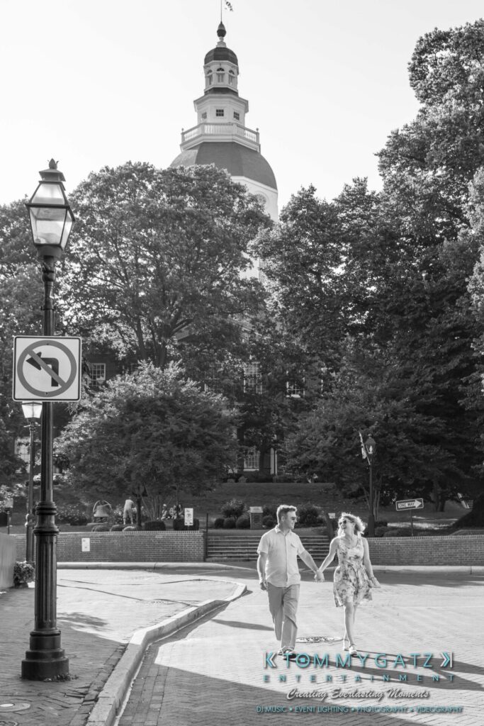 Jennifer-Rudi-Engagement-6102017-Blog-WM-35 - TOMMY GATZ ENTERTAINMENT A couple holding hands walks across a street near a large, domed building surrounded by trees.