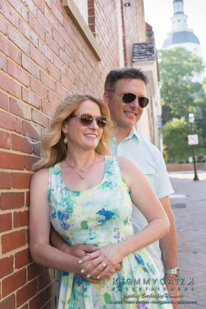 Jennifer-Rudi-Engagement-6102017-Blog-WM-37 - TOMMY GATZ ENTERTAINMENT A couple stands together in front of a brick wall. They are both wearing sunglasses and smiling. The woman is in a floral dress, and the man is in a light blue shirt.