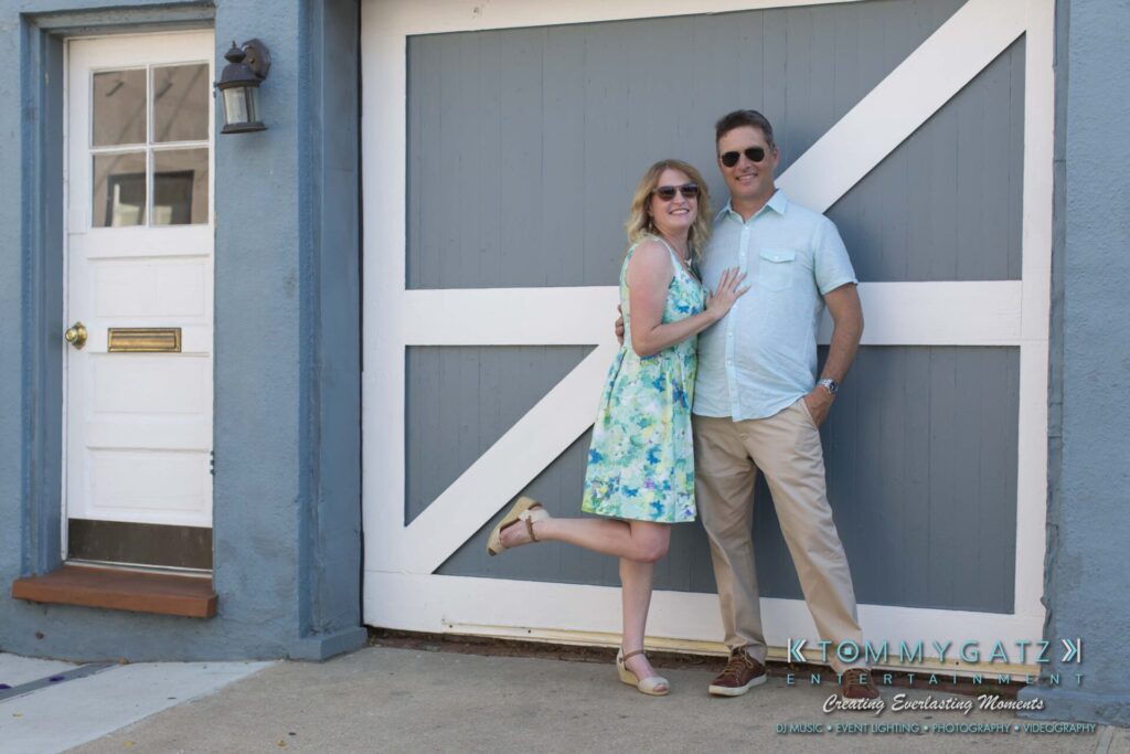 Jennifer-Rudi-Engagement-6102017-Blog-WM-4 - TOMMY GATZ ENTERTAINMENT A woman in a floral dress stands with one leg up beside a man in a light shirt and khakis, in front of a large blue and gray door.