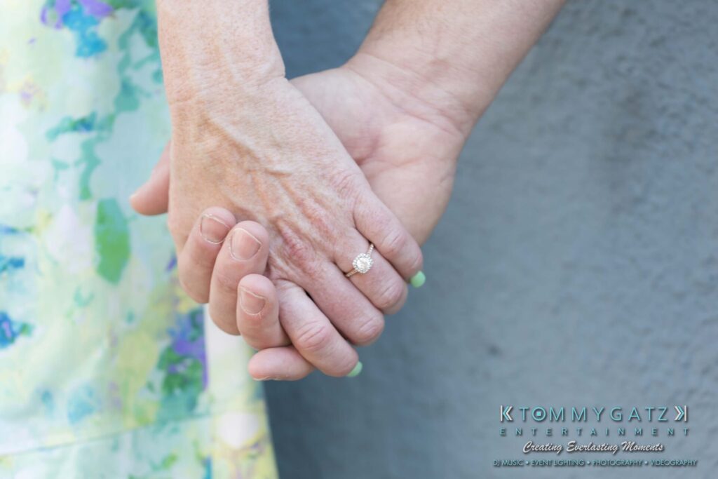 Jennifer-Rudi-Engagement-6102017-Blog-WM-40 - TOMMY GATZ ENTERTAINMENT Close-up of a couple holding hands, with one hand wearing a diamond ring. Background features floral patterns.