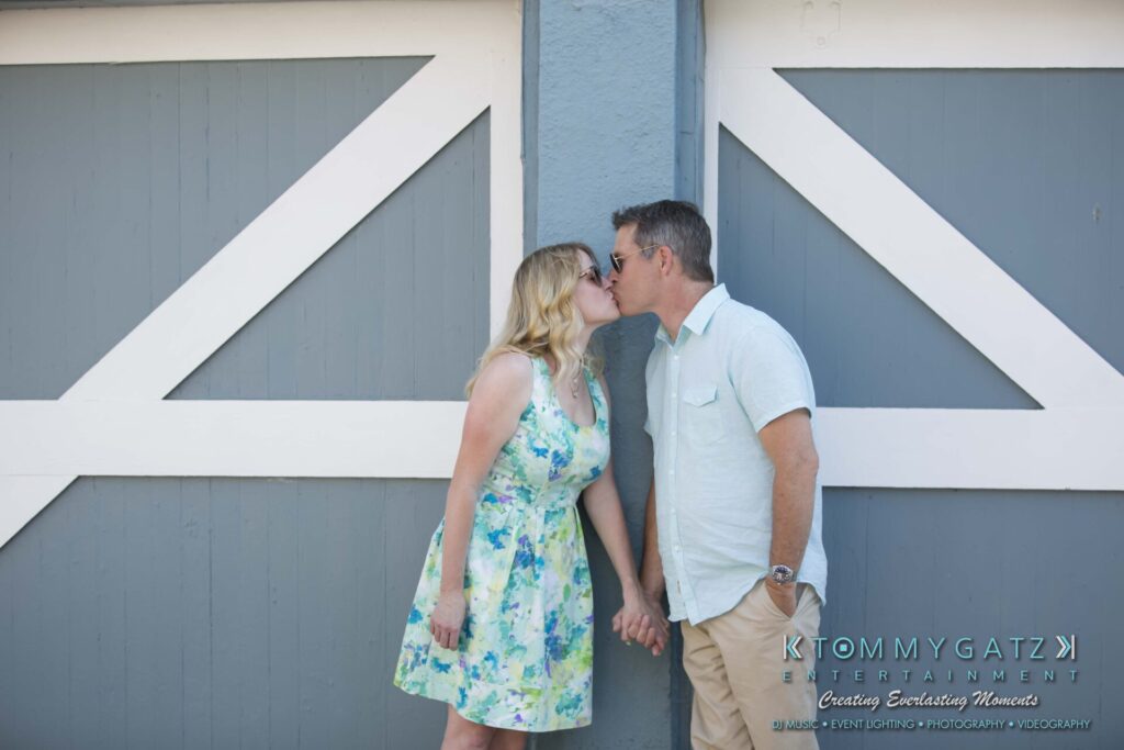 Jennifer-Rudi-Engagement-6102017-Blog-WM-42 - TOMMY GATZ ENTERTAINMENT A couple holds hands and kisses in front of a large gray and white wooden door.