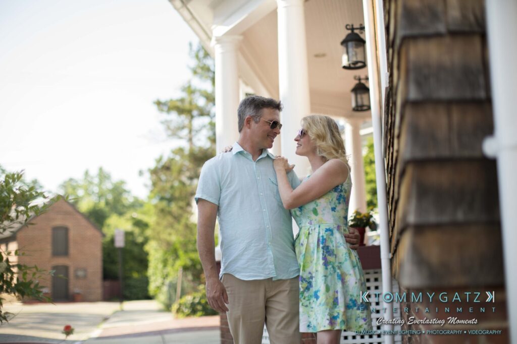 Jennifer-Rudi-Engagement-6102017-Blog-WM-45 - TOMMY GATZ ENTERTAINMENT A couple stands close on a sunny sidewalk, with the woman looking up at the man. Both wear sunglasses and light clothing. A tree and brick building are visible in the background.