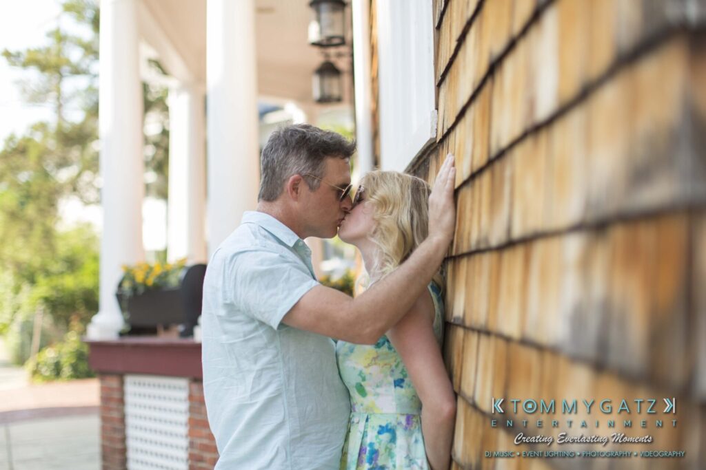 Jennifer-Rudi-Engagement-6102017-Blog-WM-47 - TOMMY GATZ ENTERTAINMENT A couple kisses in front of a house with wooden siding. Lush greenery and an intricate porch are in the background.