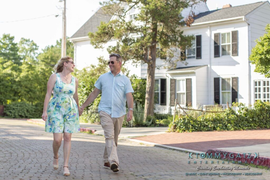Jennifer-Rudi-Engagement-6102017-Blog-WM-49 - TOMMY GATZ ENTERTAINMENT A couple walks hand in hand on a sunny day in a residential neighborhood, with a white house in the background.