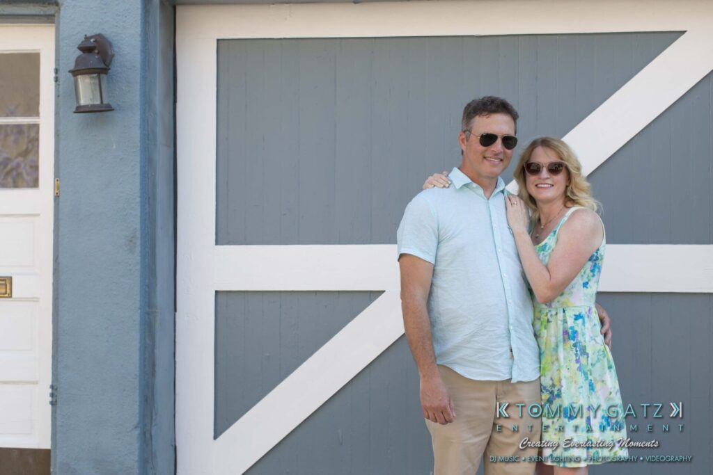 Jennifer-Rudi-Engagement-6102017-Blog-WM-5 - TOMMY GATZ ENTERTAINMENT A couple wearing sunglasses stands in front of a blue-gray garage door with white trim, smiling and embracing each other.
