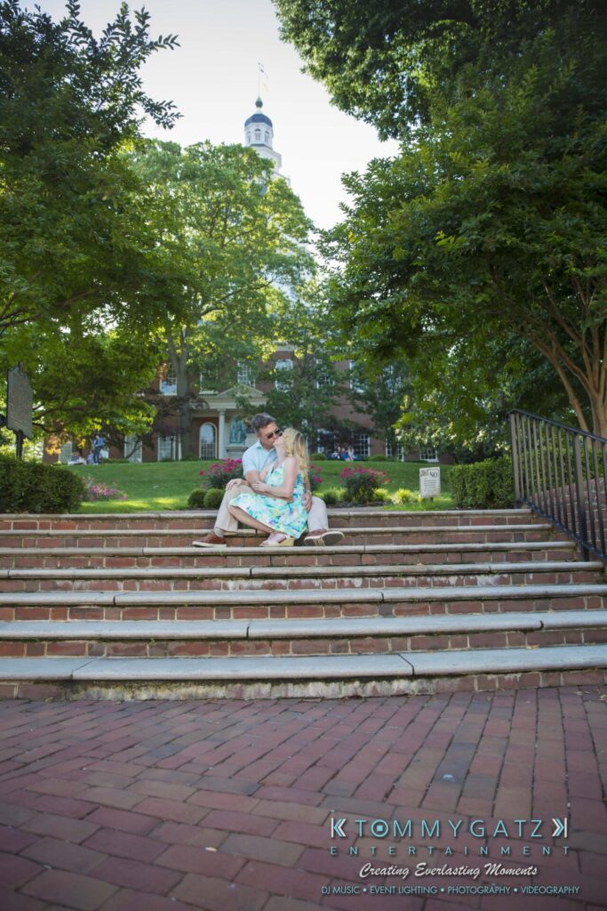 Jennifer-Rudi-Engagement-6102017-Blog-WM-50 - TOMMY GATZ ENTERTAINMENT A couple sits embracing on brick steps in a park, surrounded by trees. A building with a tower is in the background under a clear sky.