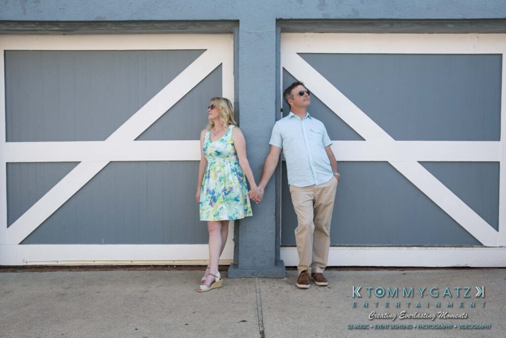Jennifer-Rudi-Engagement-6102017-Blog-WM-6 - TOMMY GATZ ENTERTAINMENT A couple stands back-to-back holding hands in front of a gray and white garage door. The woman wears a floral dress, and the man wears a light blue shirt and khaki pants.