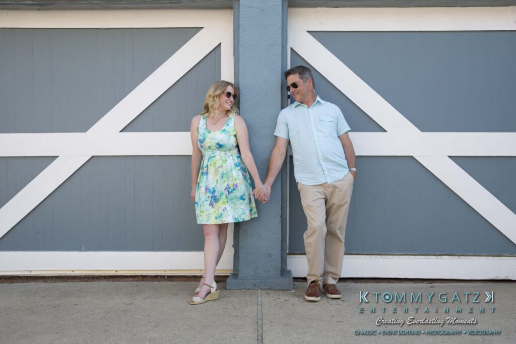 Jennifer-Rudi-Engagement-6102017-Blog-WM-7 - TOMMY GATZ ENTERTAINMENT A couple holds hands, leaning against a gray and white garage door, dressed in summer attire.
