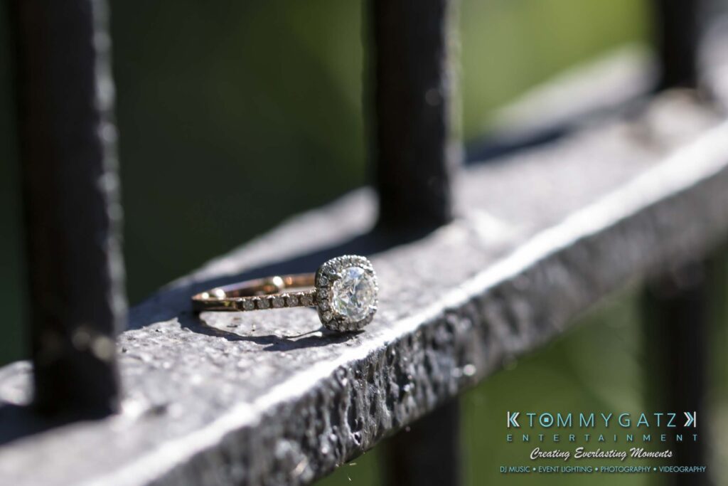 Jennifer-Rudi-Engagement-6102017-Blog-WM-9 - TOMMY GATZ ENTERTAINMENT A diamond ring rests on a metal railing in sunlight.