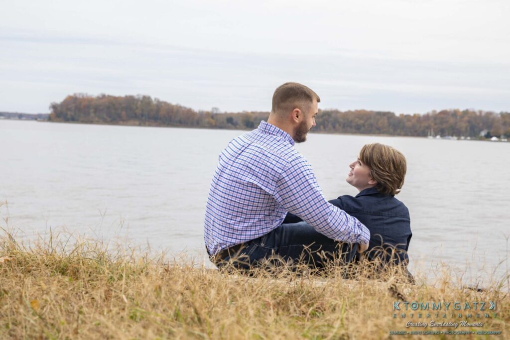 Jon-Christine-111218-Blog-Low-Res-11 - TOMMY GATZ ENTERTAINMENT Two people sitting in grass, facing a calm lake with trees on the opposite shore.