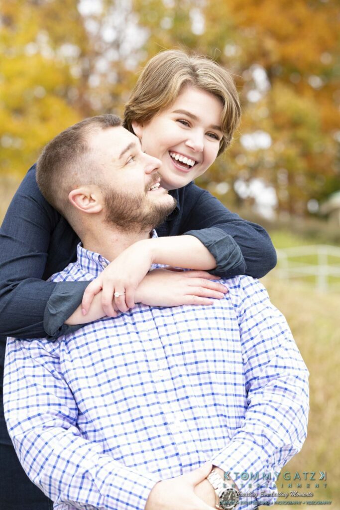 Jon-Christine-111218-Blog-Low-Res-16 - TOMMY GATZ ENTERTAINMENT A person with short hair embraces a bearded person from behind in an outdoor setting with autumn foliage. Both are smiling.