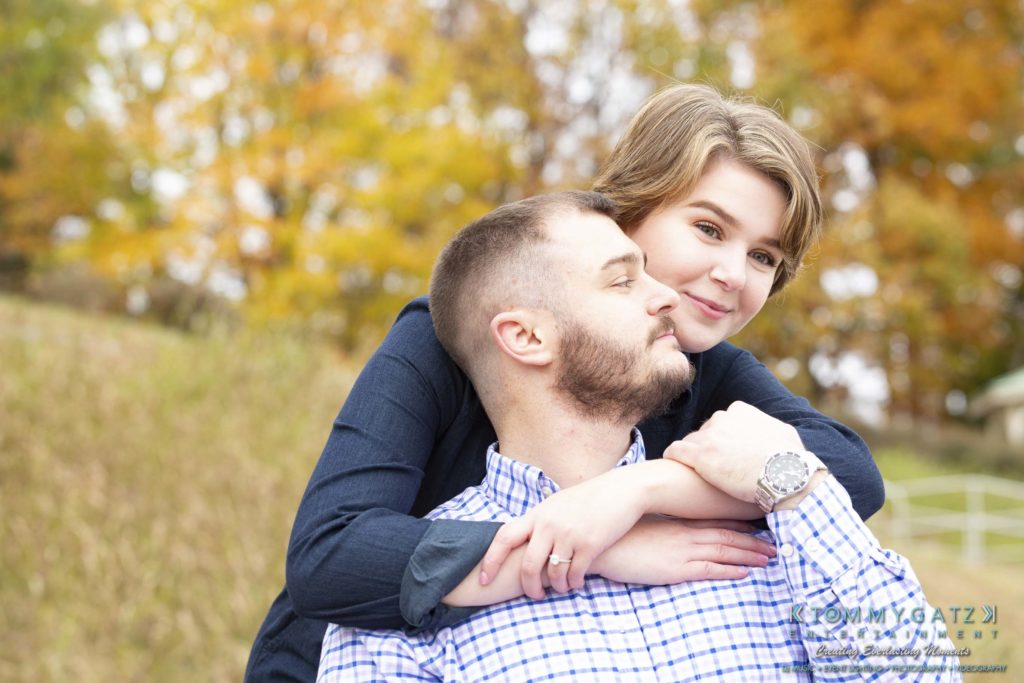 Jon-Christine-111218-Blog-Low-Res-18-1024x683 - TOMMY GATZ ENTERTAINMENT A woman embraces a man from behind in an outdoor setting with autumn foliage in the background.