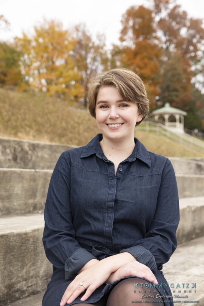 Jon-Christine-111218-Blog-Low-Res-23 - TOMMY GATZ ENTERTAINMENT A smiling person with short hair sits on outdoor steps wearing a dark dress. Autumn trees and a gazebo are in the background.