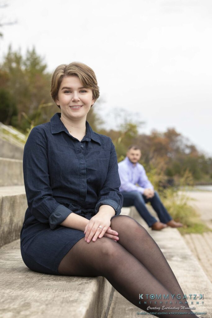 Jon-Christine-111218-Blog-Low-Res-28 - TOMMY GATZ ENTERTAINMENT A woman sits on stone steps outdoors, smiling at the camera. A man in the background sits further down the steps, looking towards the woman. Trees are visible in the distance.