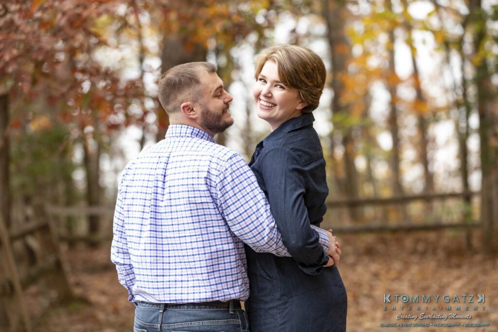 Jon-Christine-111218-Blog-Low-Res-32 - TOMMY GATZ ENTERTAINMENT A couple stands close together outdoors in a wooded area with autumn leaves. The man looks at the woman, who smiles at the camera.