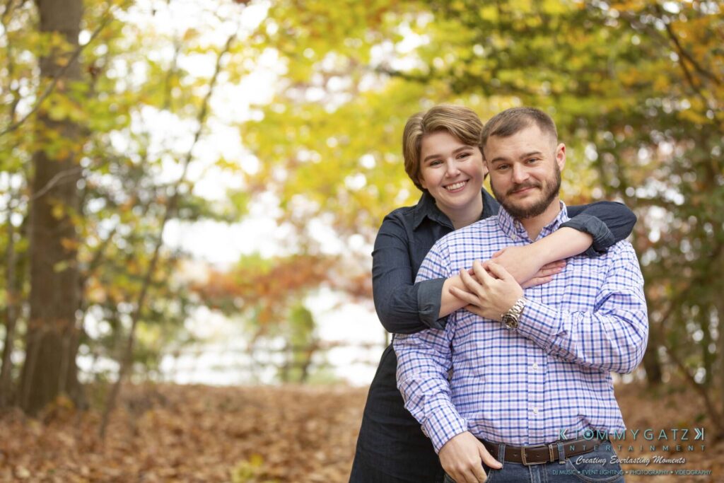 Jon-Christine-111218-Blog-Low-Res-35 - TOMMY GATZ ENTERTAINMENT A smiling couple standing in a wooded area with autumn leaves. The person behind is hugging the person in front.
