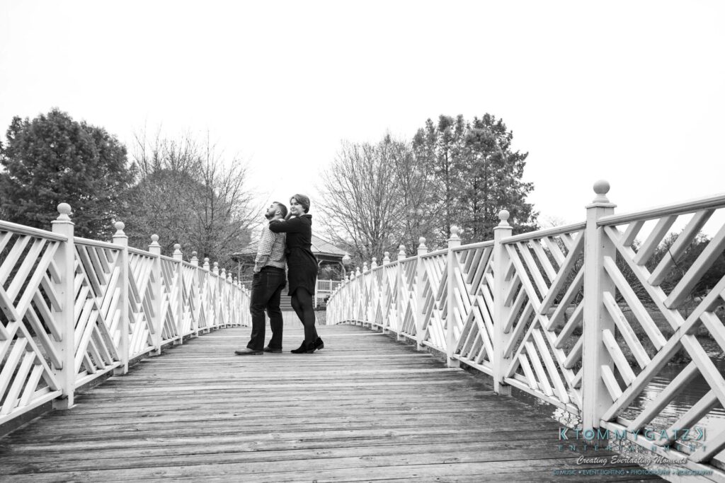 Jon-Christine-111218-Blog-Low-Res-40 - TOMMY GATZ ENTERTAINMENT Two people standing on a wooden bridge, embracing with trees in the background on a cloudy day.