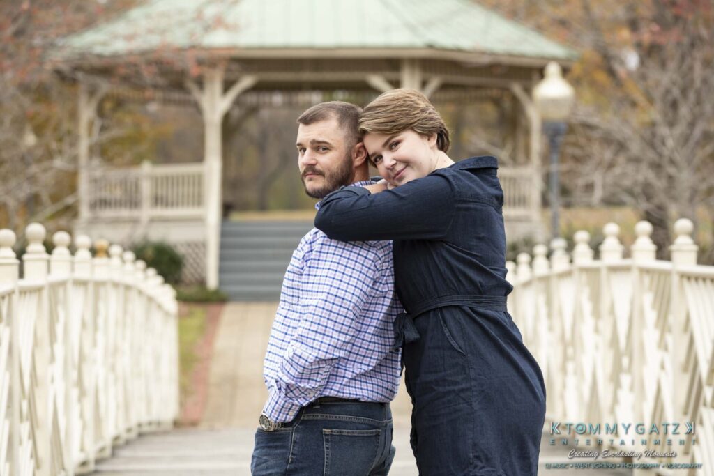 Jon-Christine-111218-Blog-Low-Res-41 - TOMMY GATZ ENTERTAINMENT A couple poses on a wooden bridge, with a gazebo and autumn trees in the background. The woman leans on the man's shoulder, both looking at the camera.