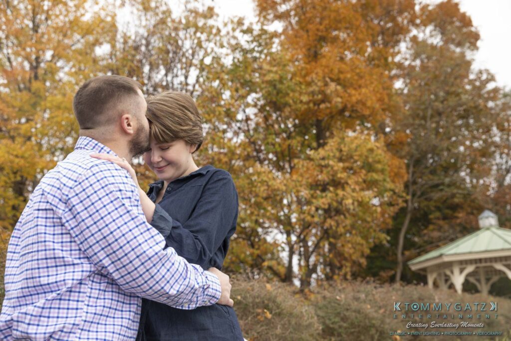 Jon-Christine-111218-Blog-Low-Res-7 - TOMMY GATZ ENTERTAINMENT A couple embraces in a park with autumn trees in the background. A small gazebo is visible to the right.