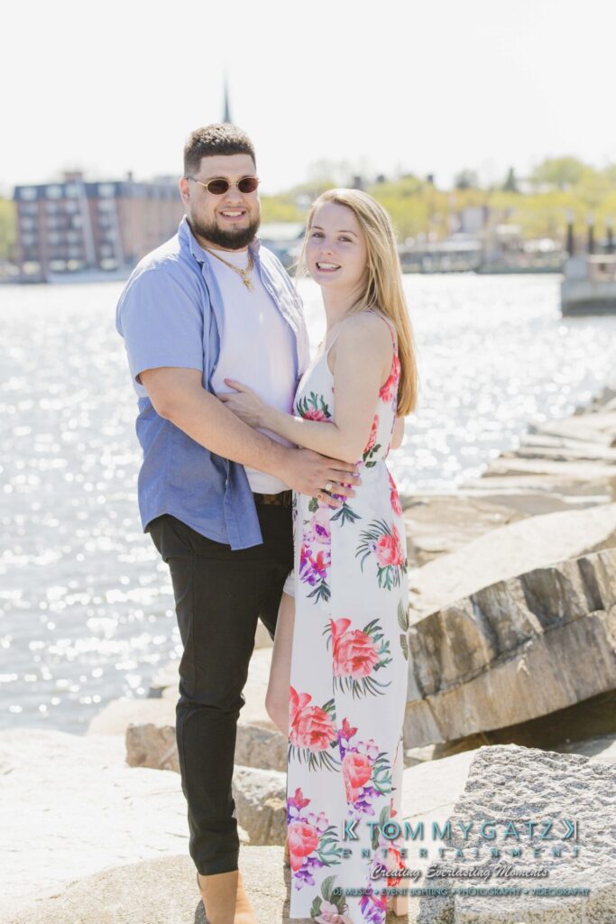 Kari-Jose-43018-FINALS-Low-Res-WM-10 - TOMMY GATZ ENTERTAINMENT A couple stands on a rocky waterfront path, with the man in a blue shirt and the woman in a floral dress, smiling at the camera.
