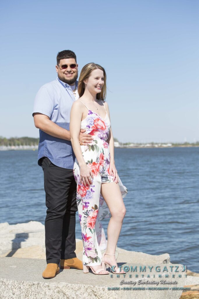 Kari-Jose-43018-FINALS-Low-Res-WM-11 - TOMMY GATZ ENTERTAINMENT A couple stands on rocks by the water. The man wears a blue shirt and black pants, and the woman wears a floral dress. They are smiling, with a sunny sky and calm water in the background.