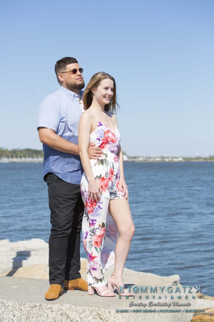 Kari-Jose-43018-FINALS-Low-Res-WM-12 - TOMMY GATZ ENTERTAINMENT A man and woman stand on rocks by the water. The man is wearing sunglasses and a blue shirt, and the woman is in a floral dress. The sky and water are clear and blue.