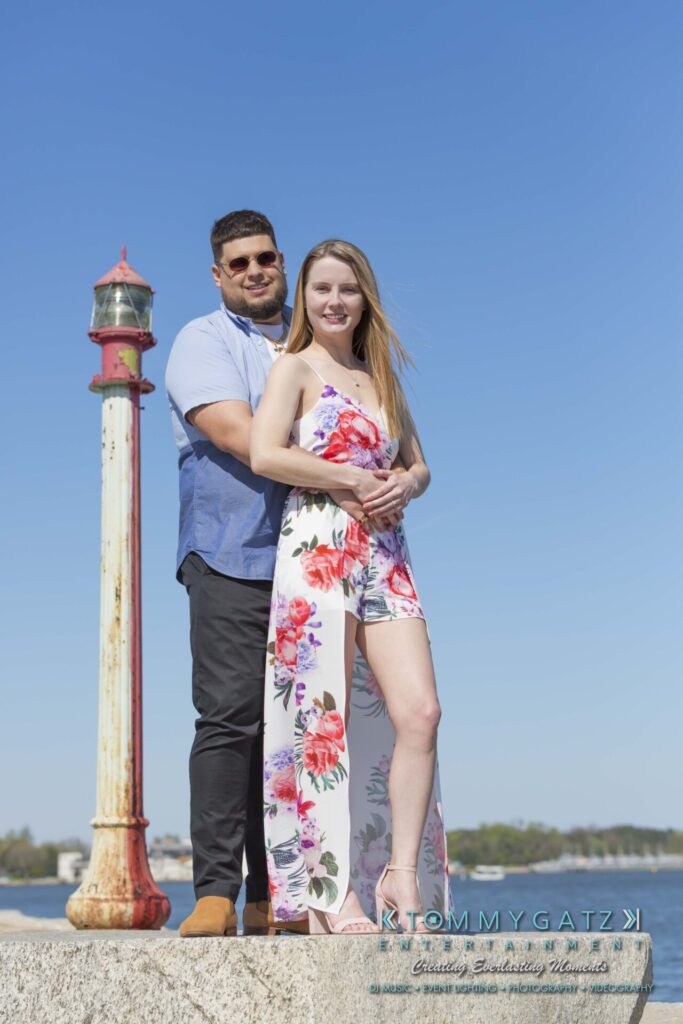 Kari-Jose-43018-FINALS-Low-Res-WM-14 - TOMMY GATZ ENTERTAINMENT A couple stands together on a pier with a red and white post in front. The woman wears a floral dress, and the man wears a blue shirt and sunglasses. A body of water is in the background.