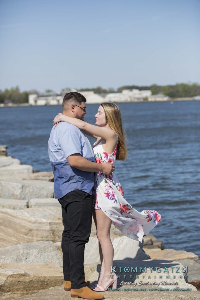 Kari-Jose-43018-FINALS-Low-Res-WM-18 - TOMMY GATZ ENTERTAINMENT A couple stands on rocks by the water, facing each other. The woman wears a floral dress, and the man wears a blue shirt.