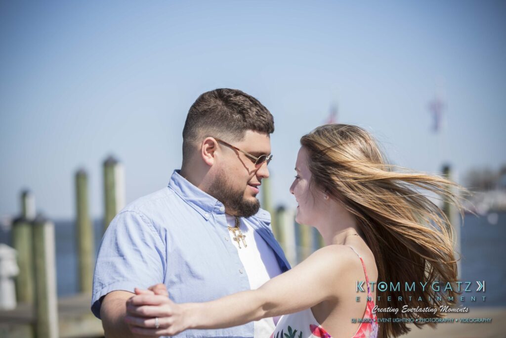 Kari-Jose-43018-FINALS-Low-Res-WM-2 - TOMMY GATZ ENTERTAINMENT A man and woman dancing outdoors near a dock. The man wears sunglasses and a blue shirt; the woman has long hair and wears a floral dress. A body of water and boats are in the background.
